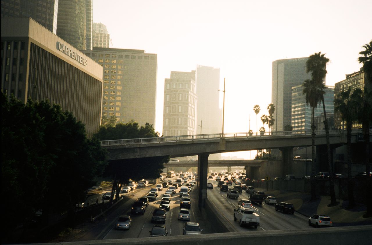 Heavy traffic on a Los Angeles freeway at sunset with buildings and palm trees in view.