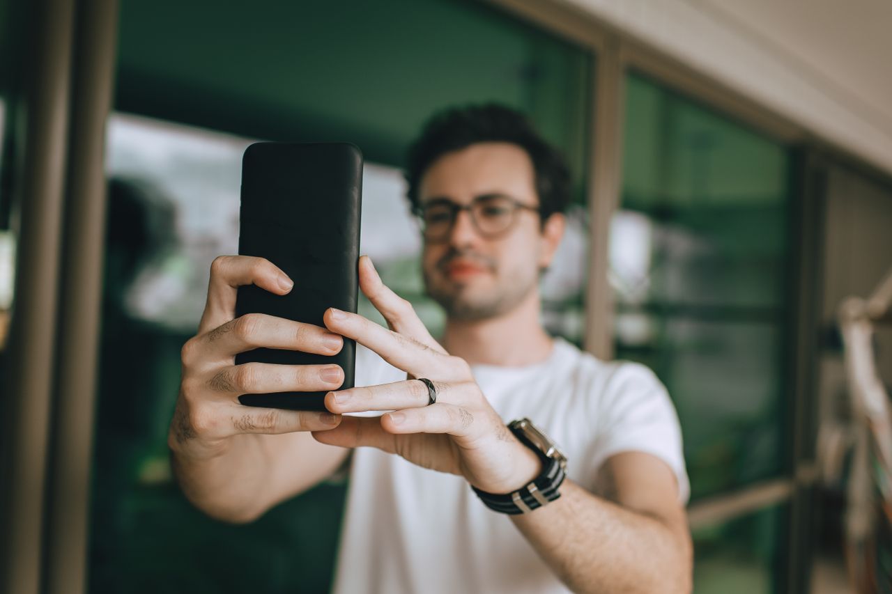 A young man takes a selfie on a balcony, using a mobile phone.