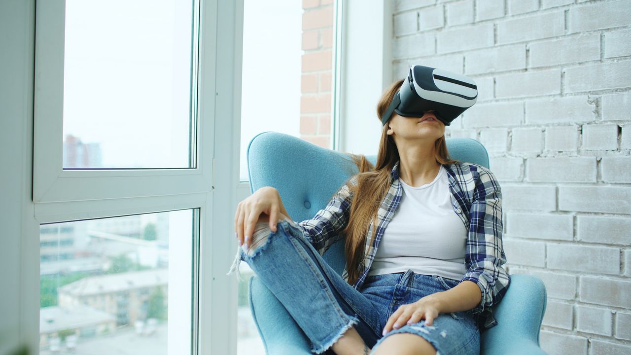Casual woman wearing VR headset sitting in a modern room looking relaxed and engaged in virtual reality.